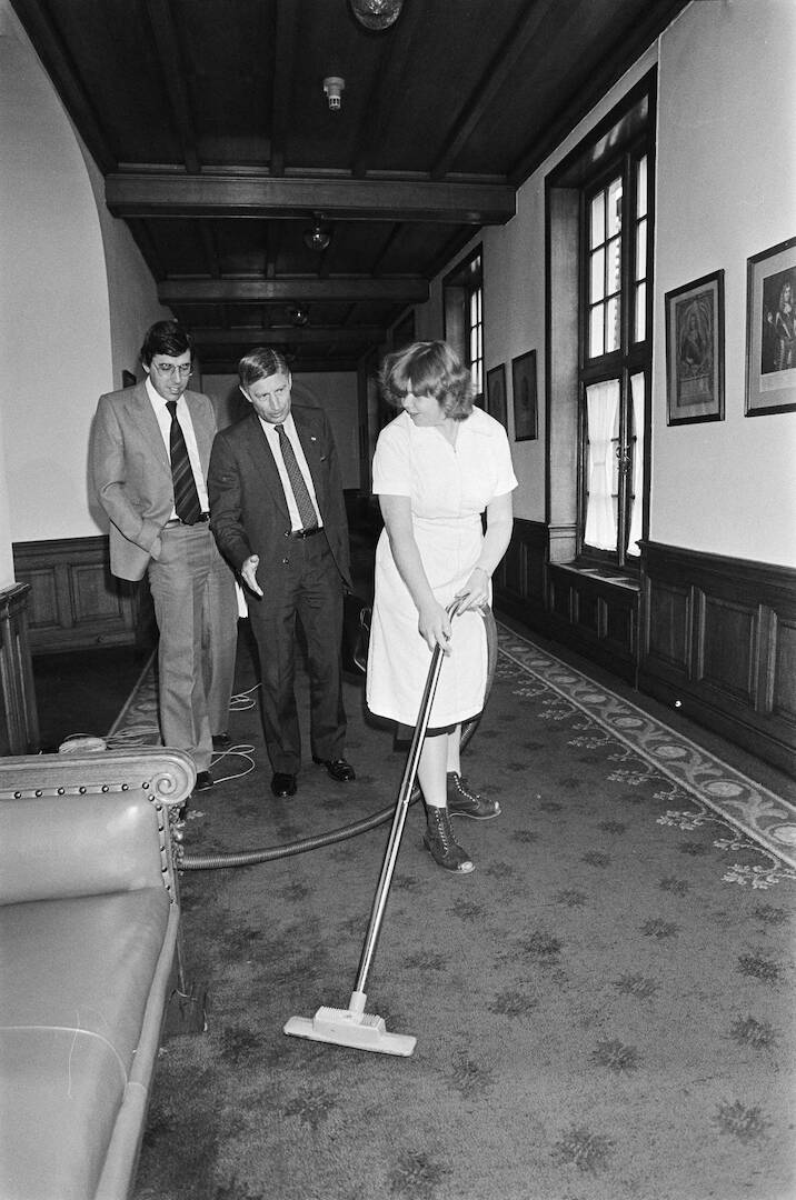 Zwart-wit foto met daarop twee mannen in gesprek met een vrouw met een stofzuiger in een gang van de Eerste Kamer