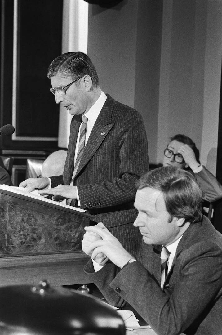 Zwart-wit foto met daarop een man staand achter het katheder in de plenaire zaal van de Eerste Kamer, een man zittend op de voorgrond