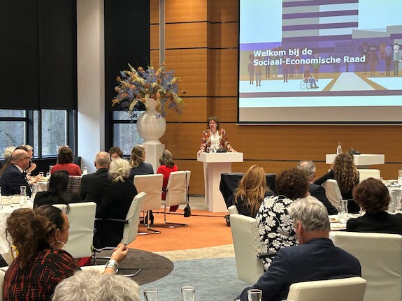 Foto van een zaal met ronde tafels waaraan mensen zitten die luisteren naar een spreker achter het spreekgestoelte. Achter haar een groot scherm met daarop de tekst "Welkom bij de SER"